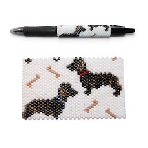 May include: A black pen with a beaded dachshund design and a matching beaded rectangle. The rectangle features two dachshunds and bone shapes on a white background. The dachshunds have black and brown fur with red and blue collars.