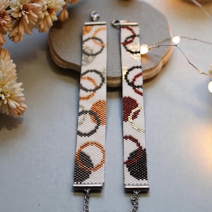 May include: Two beaded bracelets with white backgrounds and colorful ring patterns. One bracelet features orange, silver, and black rings, while the other has burgundy, gold, and black rings. Each bracelet has a silver clasp and chain.