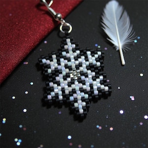 Peut inclure: Boucle d'oreille flocon de neige perlée avec un crochet argenté. Le flocon de neige est composé de perles noires, blanches et irisées. Une plume blanche est posée à côté sur une surface noire avec des paillettes.