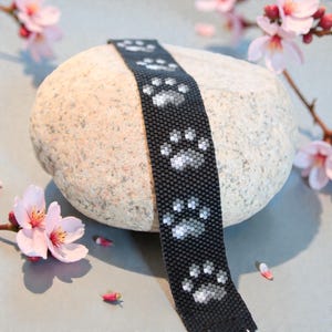 Pode incluir: Pulseira de contas pretas com pegadas brancas. A pulseira é enrolada em uma pedra clara, com flores de cerejeira rosa ao fundo. Um acessório único para os amantes de animais.