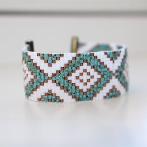 Op de afbeelding: Brede armband met een geometrisch patroon in wit, turquoise en koperkleuren. De armband heeft een metalen sluiting. Het ontwerp heeft een herhalend ruitpatroon.