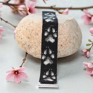Könnte beinhalten: Ein schwarzes Perlenarmband mit weißen Pfotenabdrücken. Das Armband wird auf einem glatten, hellfarbigen Stein mit rosa Blüten im Hintergrund präsentiert. Es hat einen silberfarbenen Verschluss.