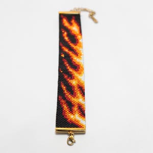 Könnte beinhalten: Ein Perlenarmband mit einem Flammendesign. Das Armband hat einen schwarzen Hintergrund mit orangefarbenen, gelben und weißen Perlen, die wie Flammen angeordnet sind. Das Armband hat goldfarbene Metallenden und einen Verschluss.