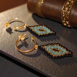 May include: Gold hoop earrings featuring diamond-shaped pendants with turquoise and black beading. The earrings are presented on a dark surface, with a leather-bound book visible. The earrings have a post and clasp.