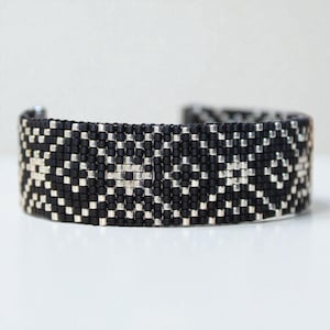 May include: A black and silver beaded cuff bracelet. The bracelet features a geometric pattern of small black and silver beads. The cuff is a wide band, with a slightly curved shape. The design is symmetrical and appears to be hand-crafted.