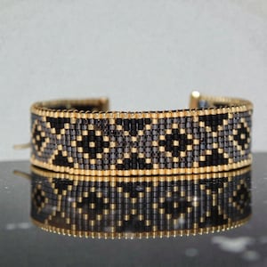 Op de afbeelding: Een kralen manchetarmband met een geometrisch patroon. De armband heeft een goudkleurige metalen basis en is versierd met kleine, gelijkmatig verdeelde kralen in zwart, grijs en goud. De armband is ongeveer 3,8 cm breed.