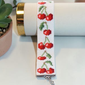 Op de afbeelding: Witte kralenarmband met een herhaald patroon van rode kersen en groene bladeren. De armband is gemaakt van kleine kralen en heeft een zilverkleurige sluiting. De armband is ongeveer 2 cm breed.