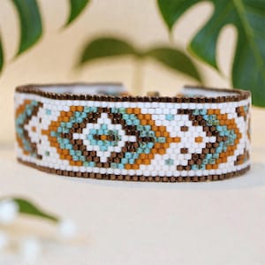 May include: A wide beaded bracelet with a geometric pattern in white, turquoise, brown, and orange. The bracelet has a brown border and is likely handmade. The design is reminiscent of Southwestern or Native American art.