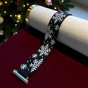 May include: A wide beaded bracelet featuring a black background with a repeating white snowflake pattern. The bracelet is constructed from small, uniform beads and is finished with a silver-toned metal clasp at one end, showcasing a winter theme.