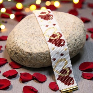 May include: White beaded bracelet with a pattern of red hearts and gold accents. The bracelet is displayed on a gray stone, surrounded by red rose petals. The bracelet has a gold clasp.