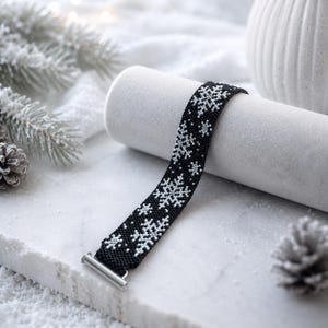 May include: A black woven bracelet featuring a repeating white snowflake pattern. The band is constructed from small beads and includes a silver-toned metal bar clasp at one end, providing a secure closure for this winter-themed accessory.
