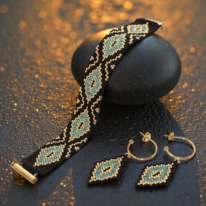 Puede incluir: Un conjunto de pulsera y pendientes a juego. La pulsera y los pendientes presentan un fondo negro con un patrón repetido de cuentas turquesas y doradas. Los pendientes son aros dorados con un colgante en forma de diamante con cuentas.