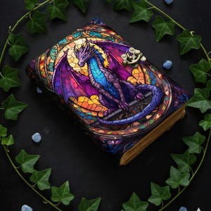Könnte beinhalten: Ein Fantasy-Buch mit einem Einband im Glasmalerei-Stil, der einen lila und blauen Drachen zeigt. Das Buch hat einen Metallverschluss und gealterte, vergilbte Seiten. Grüner Efeu und kleine blaue Steine umgeben das Buch auf einer schwarzen Oberfläche.