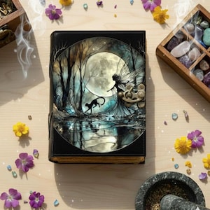 Puede incluir: Un diario encuadernado en cuero negro con una ilustración de fantasía en la portada. La obra de arte representa un hada, un gato negro y una luna llena sobre una masa de agua reflectante. Flores amarillas y moradas y cristales rodean el diario.