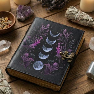 May include: Black leather journal featuring a moon phase design with purple floral accents. The journal has a metal clasp and aged paper edges. The image is set on a wooden surface with candles, crystals, and dried herbs.