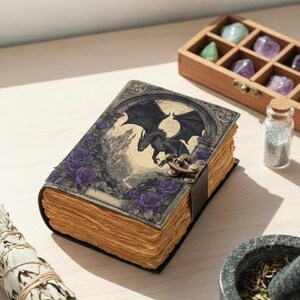 May include: A fantasy journal with a dragon illustration on the cover, featuring purple roses and a castle backdrop. The journal has a clasp closure and aged pages. A wooden box with crystals, a small glass bottle, and a mortar and pestle are also visible.
