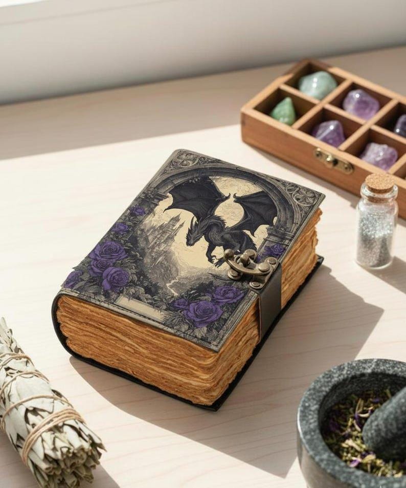 May include: A fantasy journal with a dragon illustration on the cover, featuring purple roses and a castle backdrop. The journal has a clasp closure and aged pages. A wooden box with crystals, a small glass bottle, and a mortar and pestle are also visible.