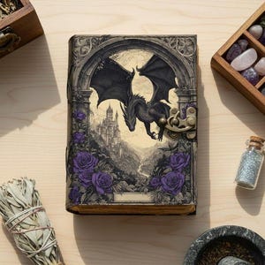 May include: A fantasy journal with a dragon illustration. The cover depicts a black dragon with outstretched wings, a castle, and purple roses. The journal has a clasp closure and is surrounded by other items.