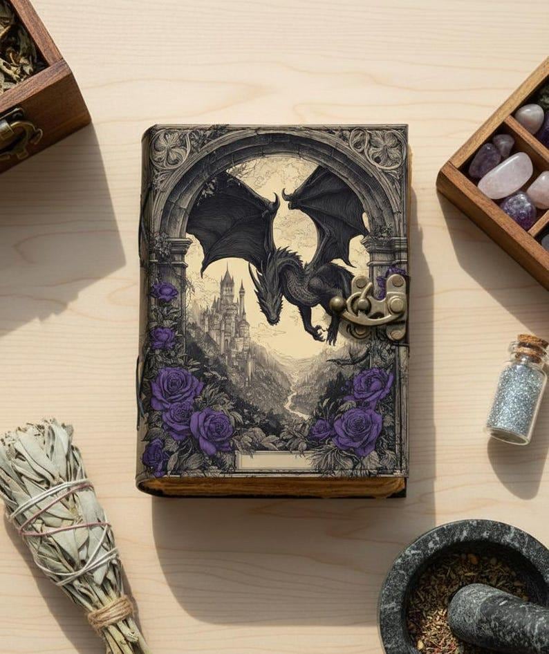 May include: A fantasy journal with a dragon illustration. The cover depicts a black dragon with outstretched wings, a castle, and purple roses. The journal has a clasp closure and is surrounded by other items.