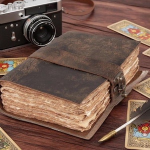Puede incluir: Un diario de cuero marrón con un cierre y una correa de metal, sobre una superficie de madera. También hay una cámara vintage y cartas del tarot. Las páginas del diario tienen un aspecto rústico y envejecido.