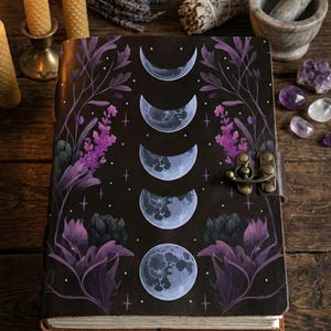 May include: A dark brown journal with a moon phase design, featuring a series of crescent moons transitioning to a full moon. The cover is adorned with purple floral accents and a metal clasp. Candles and crystals are in the background.