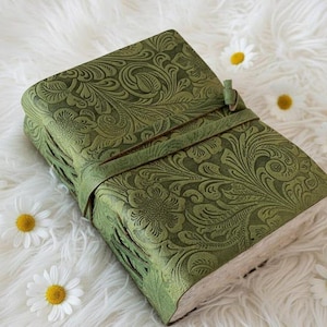 May include: A green leather journal with an embossed floral pattern. The journal features a strap closure and is placed on a white, fluffy surface. The pages' edges are visible.