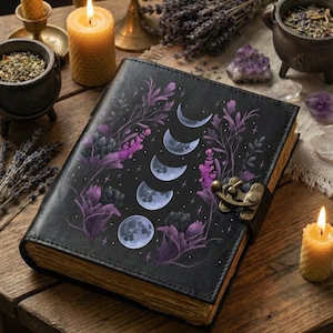 May include: A black leather-bound journal with a moon phase design in purple and white. The journal is closed with a metal clasp. The image includes lit candles, herbs, and crystals, creating a mystical aesthetic.