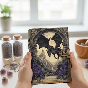May include: A fantasy journal featuring a black dragon illustration. The cover depicts a dragon flying over a castle, framed by purple roses. The journal has a clasp closure and a leather-like texture.