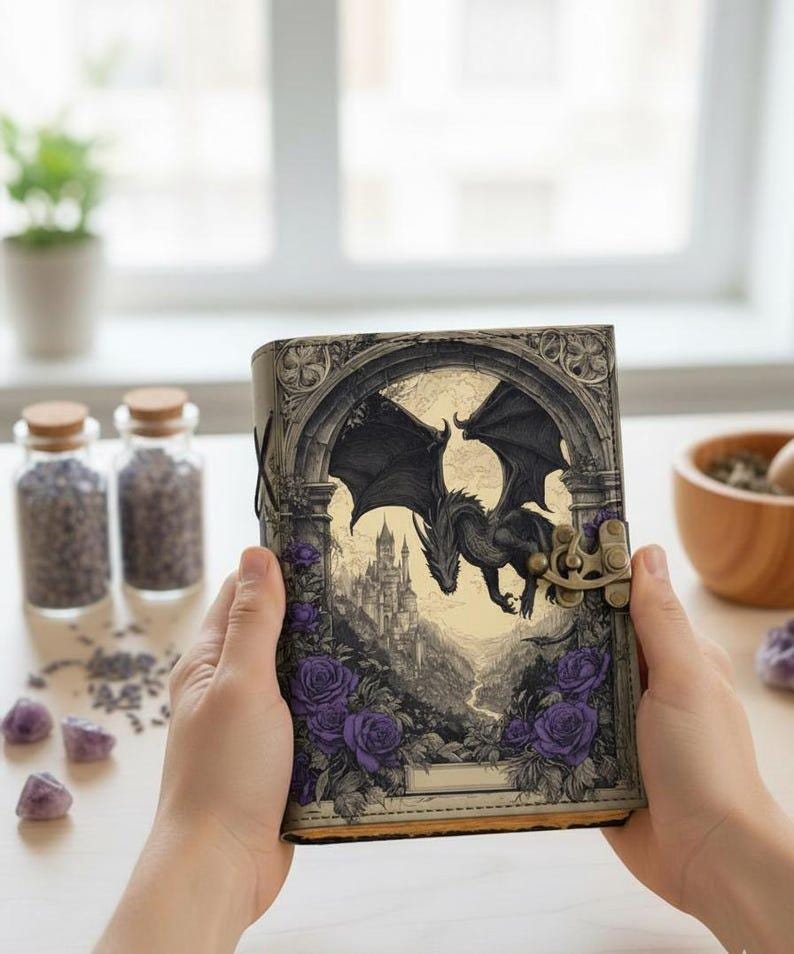 May include: A fantasy journal featuring a black dragon illustration. The cover depicts a dragon flying over a castle, framed by purple roses. The journal has a clasp closure and a leather-like texture.