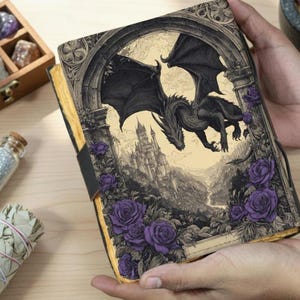 May include: A fantasy journal with a black dragon illustration. The cover shows a detailed drawing of a dragon flying over a castle, framed by purple roses. The journal has a black elastic band closure.