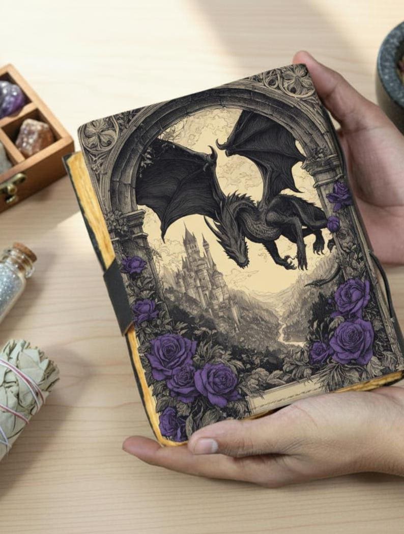 May include: A fantasy journal with a black dragon illustration. The cover shows a detailed drawing of a dragon flying over a castle, framed by purple roses. The journal has a black elastic band closure.