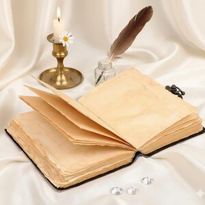 May include: An open antique book with aged, yellowed pages, a black cover, and a metal clasp. A lit candle in a brass holder, a feather quill, and an inkwell sit nearby. Three clear, sparkling gems are at the bottom.