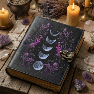May include: Black leather journal with a moon phase design in purple and white. The journal is closed with a metal clasp. The background includes candles, crystals, and a cauldron, suggesting a mystical or witchy theme.