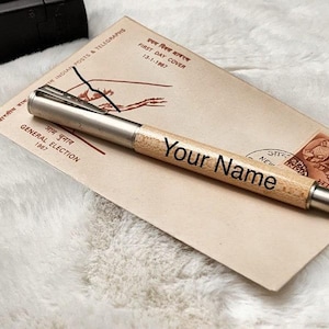 May include: A personalized pen with "Your Name" printed on the wooden barrel, resting on a vintage envelope. The pen has a silver metal tip and clip. The envelope has text in English and Hindi, with a stamp.