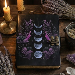 May include: Black journal with a moon phase design, purple floral accents, and a metal clasp. The journal is open on a wooden table with candles, dried lavender, and crystals. The cover features a series of moon phases.