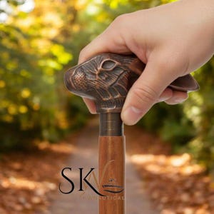Può includere: Bastone da passeggio con manico a forma di testa di coniglio in bronzo. L'asta è in legno e presenta un collare in metallo. Lo sfondo sfocato mostra un sentiero e vegetazione. Il logo "SK Nautical" è visibile in basso.
