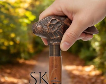 May include: A bronze-colored walking cane with a rabbit head handle. The cane has a wooden shaft and a metal collar. The image is set against a blurred background of a path and foliage. The logo "SK Nautical" is at the bottom.