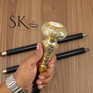 Puede incluir: Bastón de color latón con un mango decorativo con una esfera de reloj. El bastón tiene un eje negro y se sostiene en una mano. El logotipo "SKWNautical" es visible en el fondo.