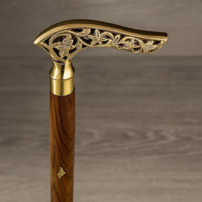 May include: A decorative walking cane with a brass handle featuring a floral design. The handle curves downward and is attached to a wooden shaft. The shaft has a small, gold-colored inlay near the bottom.
