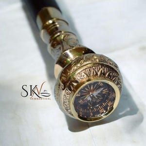 May include: Close-up of a brass walking cane handle with an embedded compass. The handle is gold-colored with intricate engraved patterns. The compass face is black with gold markings. The cane's shaft is dark brown.