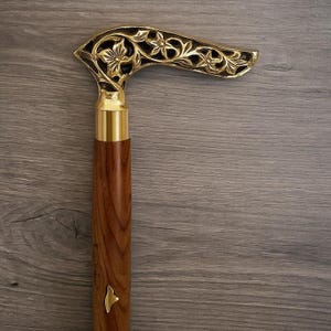 May include: A wooden walking cane with a decorative gold-colored handle. The handle features an intricate floral design. The shaft is a rich brown color, and the handle is accented with gold-colored metal.