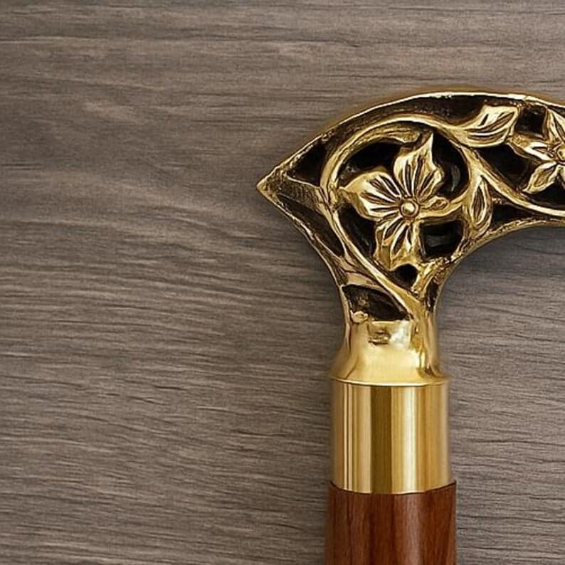 May include: Close-up of a decorative walking cane handle. The handle is made of gold-colored metal with a floral design. The lower portion of the handle is a polished brass color, and the shaft is a dark brown wood.