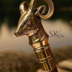 May include: A brass walking cane with a ram's head design. The ram's head is detailed with curved horns and a textured surface. The handle is made of dark wood and has brass accents. The cane has the text "SK" on it.