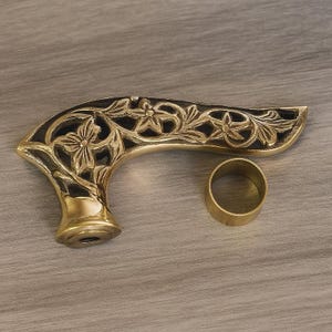 May include: A decorative brass handle with an intricate floral design and a separate brass ring. The handle has a curved shape and a dark background, with the floral details standing out in a gold color.