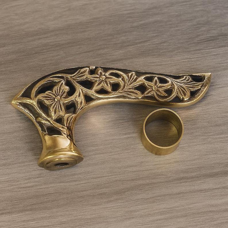 May include: A decorative brass handle with an intricate floral design and a separate brass ring. The handle has a curved shape and a dark background, with the floral details standing out in a gold color.