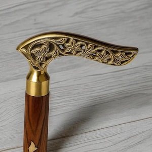 Könnte beinhalten: Ein Gehstock aus Holz mit einem Messinggriff. Der Griff ist aufwendig mit Blumen- und Blättermustern verziert. Ein Messingkragen verbindet den Griff mit dem Holzschaft. Ein klassisches Accessoire.