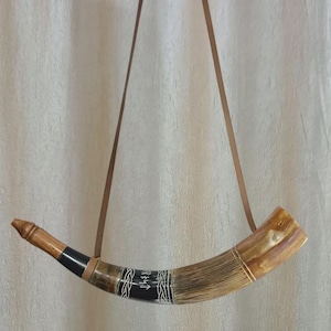 Könnte beinhalten: Ein handgefertigtes Horn, wahrscheinlich zum Trinken oder für zeremonielle Zwecke, mit einem braunen Lederriemen. Das Horn hat eine hellbraune und beige Farbgebung mit schwarzen und weißen Zierbändern. Das Horn ist etwa 30 cm lang.
