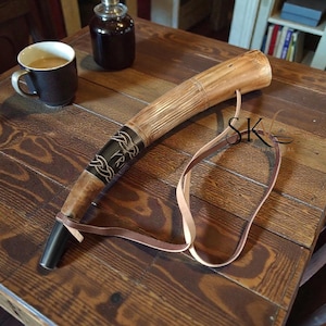 Könnte beinhalten: Ein handgefertigtes Trinkhorn mit Lederriemen, das auf einem Holztisch liegt. Das Horn ist hellbraun mit einem dunklen Band mit einem komplizierten Design. Eine Tasse und eine Flasche befinden sich ebenfalls auf dem Tisch.