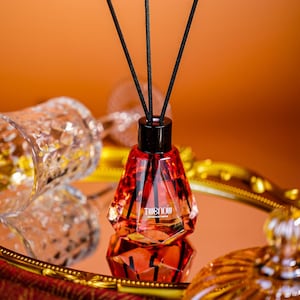 May include: A decorative red and black essential oil diffuser with black reeds. The diffuser has a geometric design and the text "TWENOW" is visible on the front. The diffuser sits on a gold-rimmed mirror.