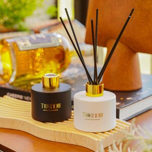 Może przedstawiać: Dwa ceramiczne dyfuzory zapachowe z patyczkami, jeden czarny i jeden biały, ze złotymi akcentami i czarnymi patyczkami. Dyfuzory stoją na drewnianej tacy. Tekst "Twenow Diffuseur de Parfum" jest wydrukowany z przodu każdego dyfuzora.